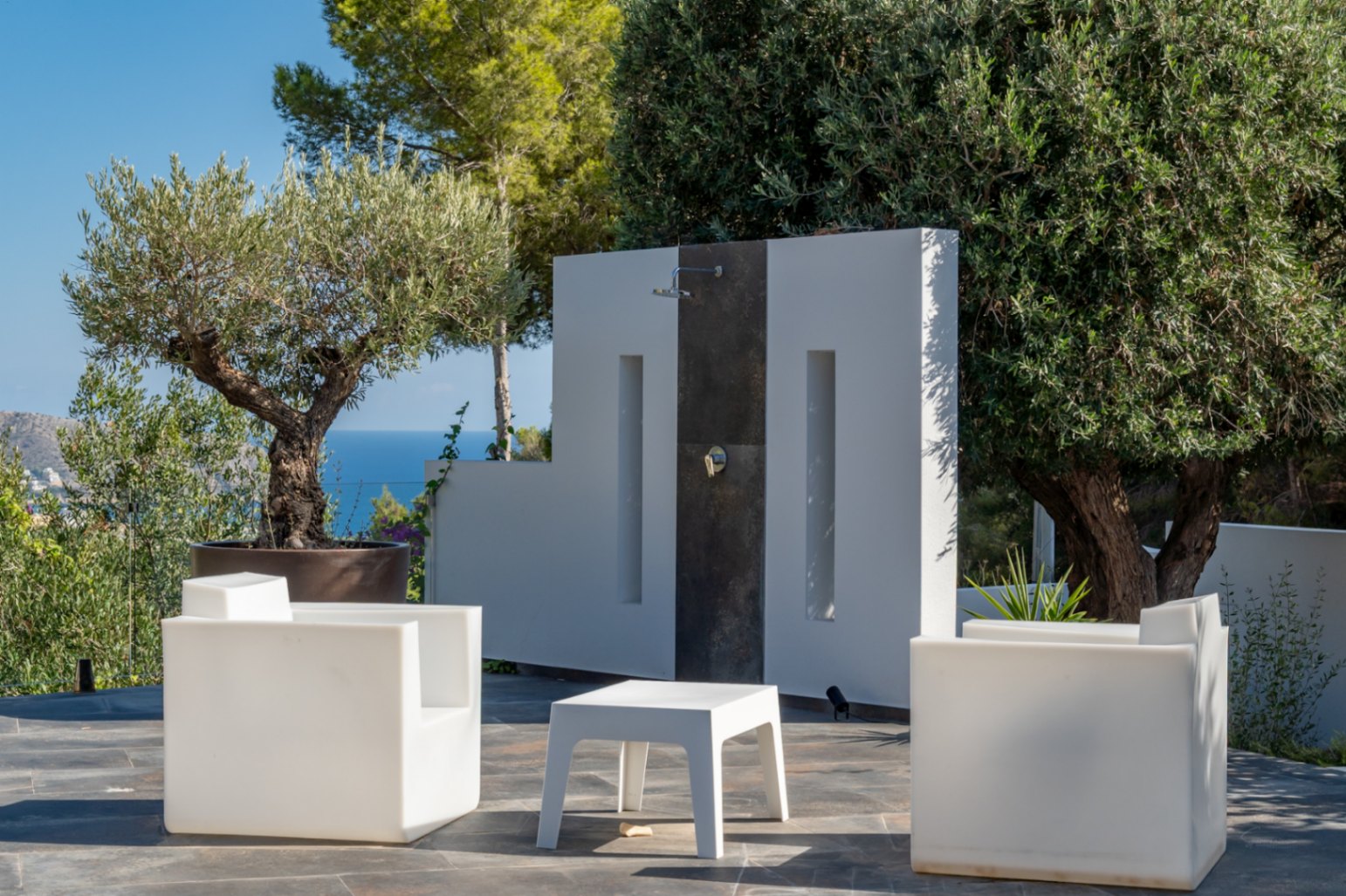 Minimalist outdoor shower area featuring white designer furniture, slate flooring, and a backdrop of the Mediterranean Sea and lush greenery.