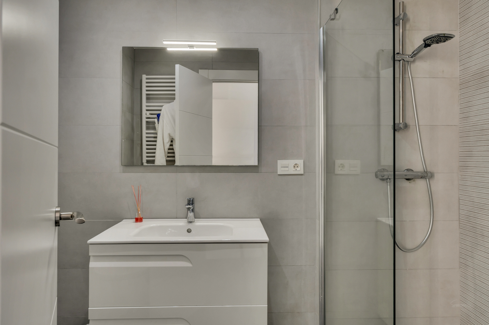 Modern bathroom featuring grey tiling, a white vanity unit, a mirror with integrated lighting, and a glass-enclosed walk-in shower with chrome fixtures.
