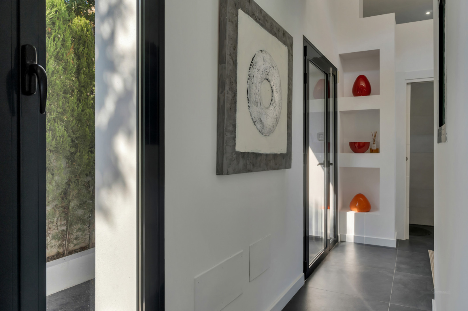 Modern hallway featuring dark porcelain flooring, built-in decorative niches, and black-framed windows that provide abundant natural light and garden views.