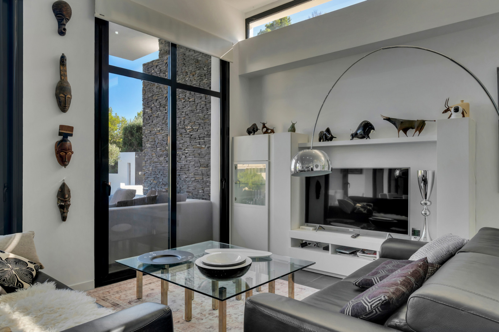 Modern living space featuring high ceilings, floor-to-ceiling glass doors, and a minimalist media unit with a chrome arc lamp and ethnic art.