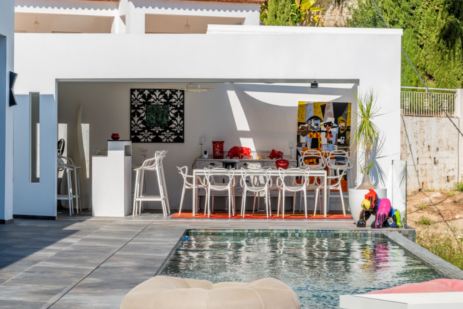 Modern summer kitchen and outdoor dining area adjacent to the pool, featuring contemporary artwork, designer white seating, and large-format grey tiling.
