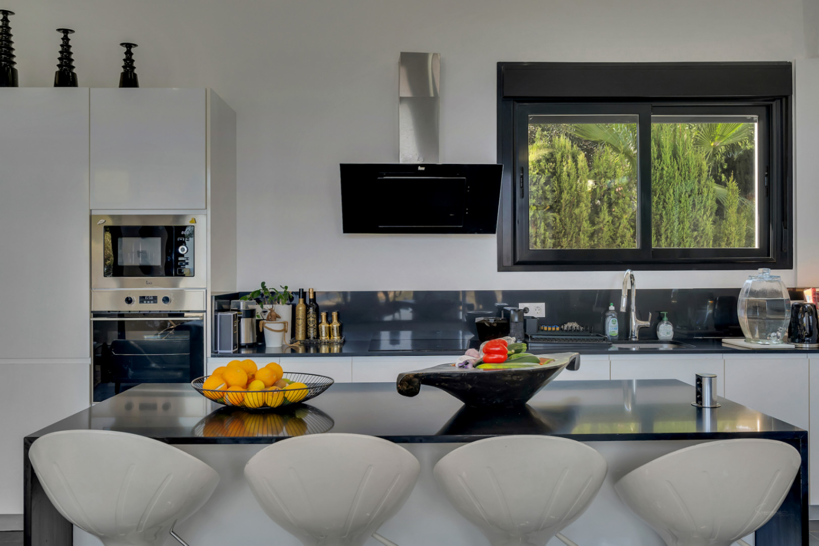 Open-concept kitchen featuring a black island with designer stools and a window overlooking lush Mediterranean greenery.