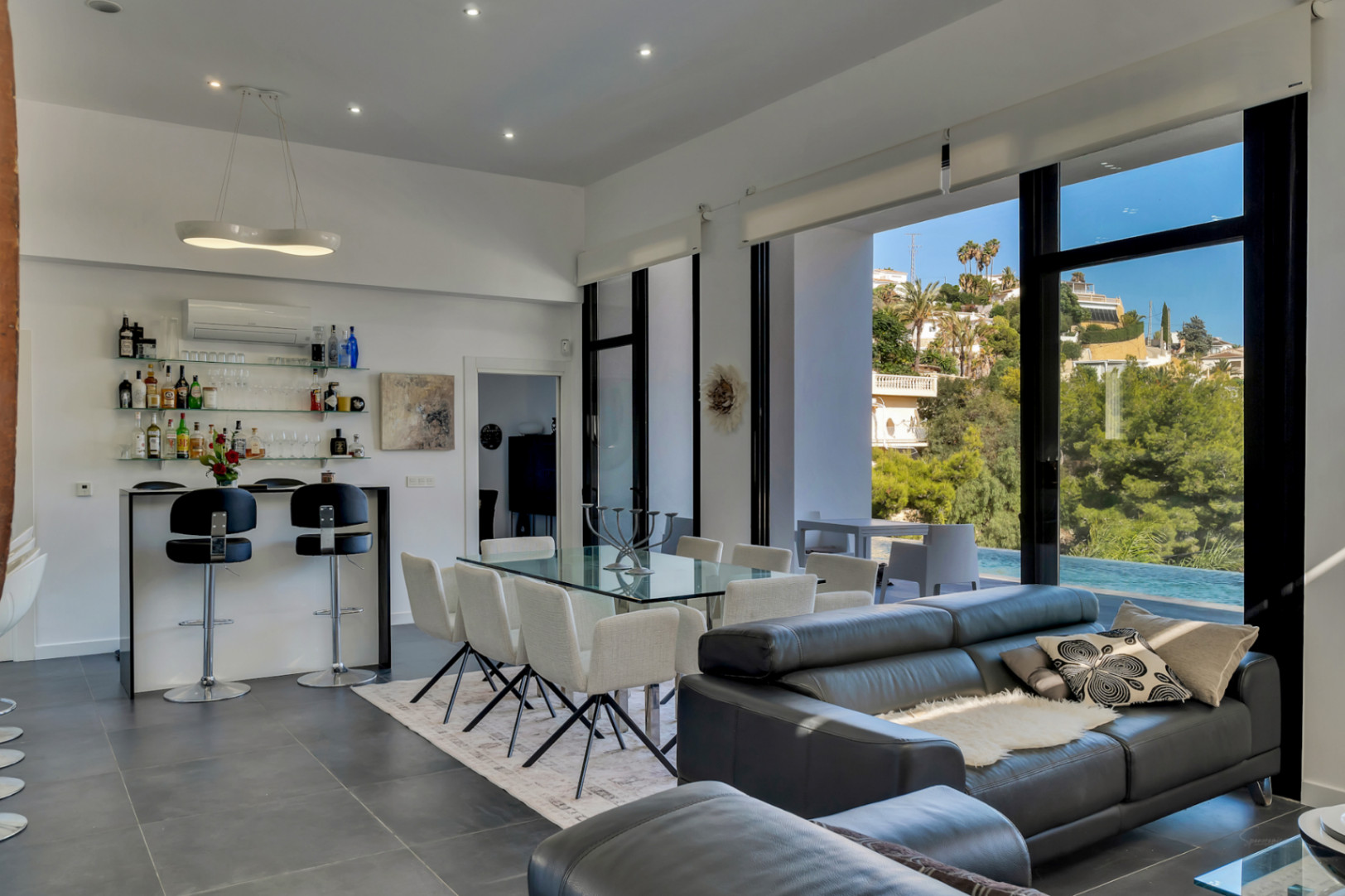 Open-plan living and dining area featuring high ceilings, a built-in bar, and floor-to-ceiling windows opening onto the pool terrace with coastal views.