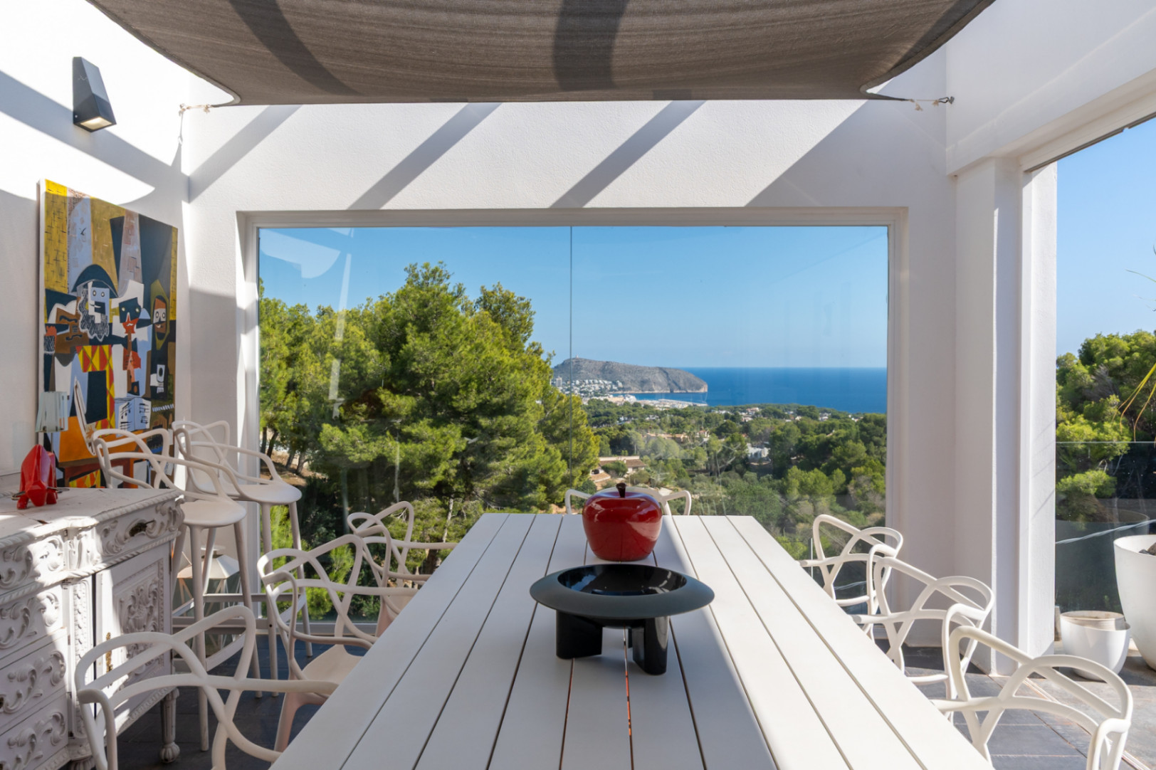Outdoor dining area featuring a white slatted table and designer chairs, sheltered by an awning with panoramic views of the Mediterranean Sea and pine forests.
