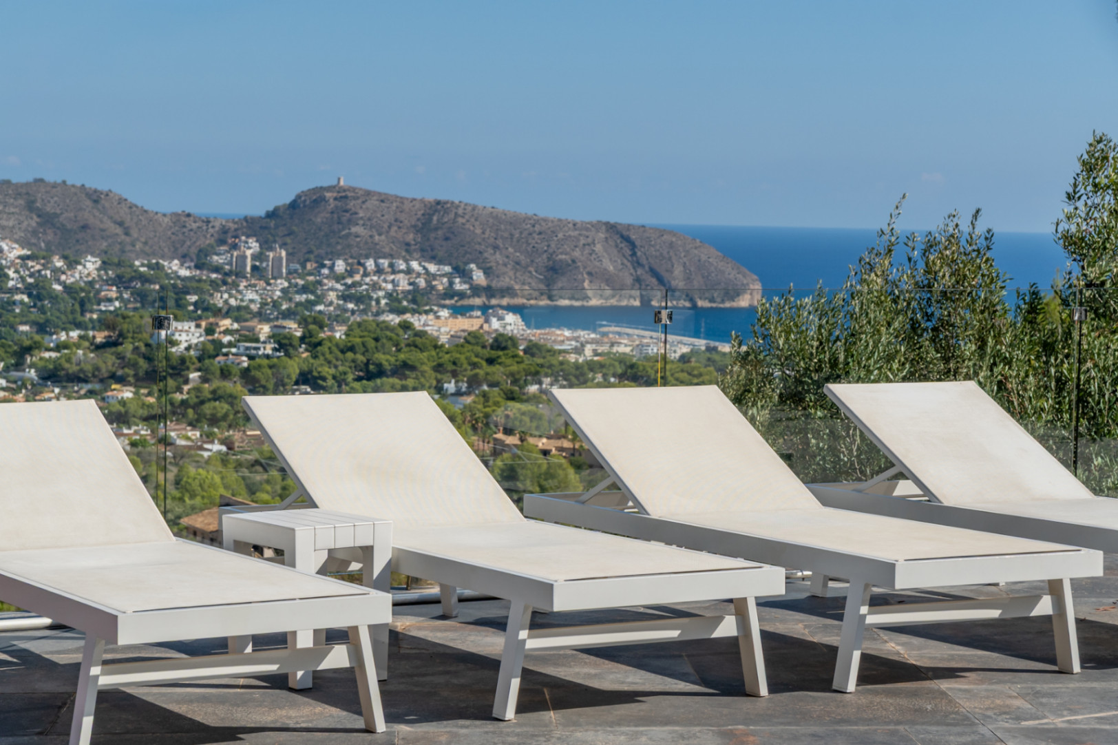 Outdoor solarium with modern white sun loungers and glass railings, offering panoramic views of the Mediterranean Sea and Moraira's coastline.
