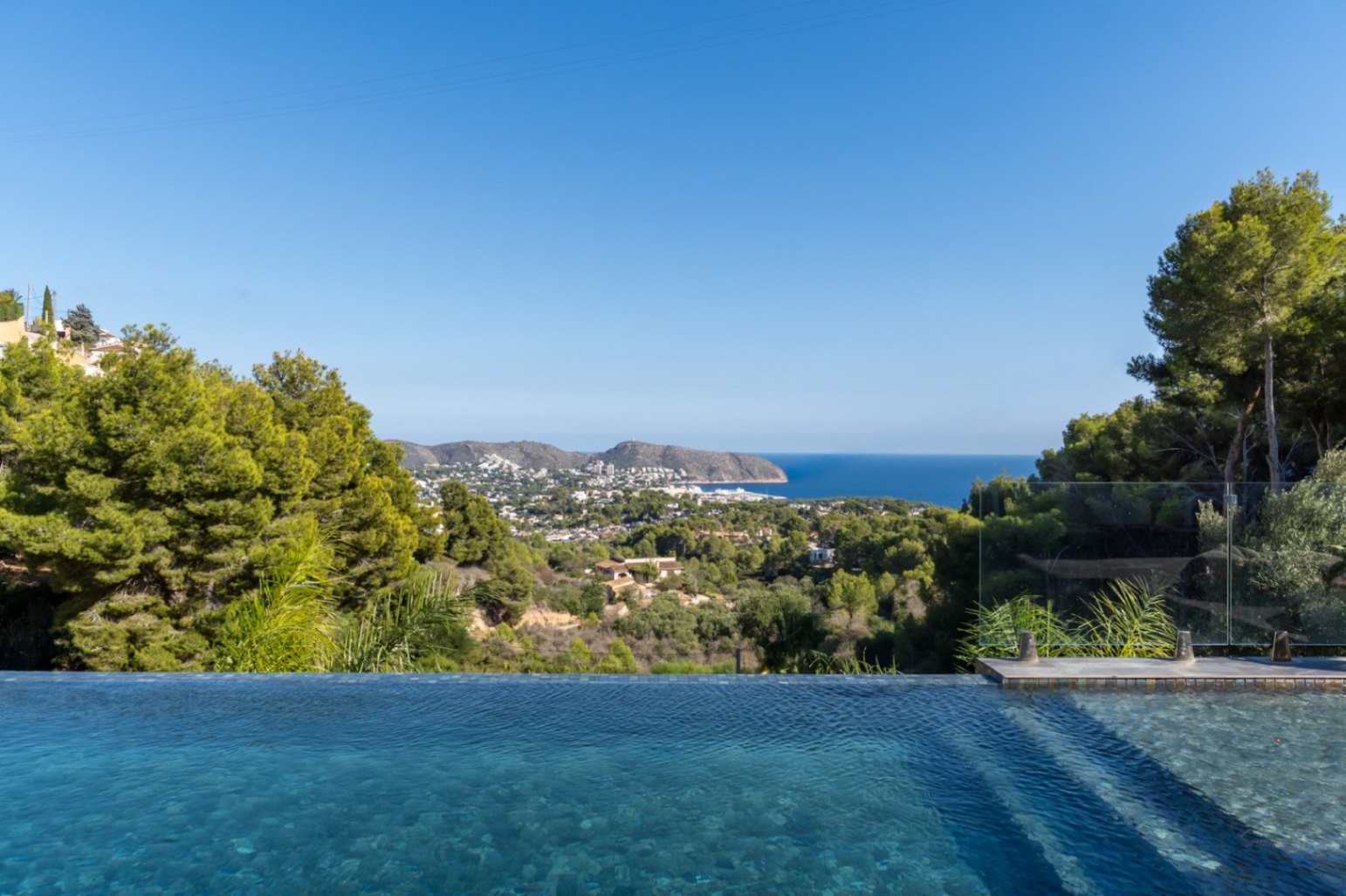 Panoramic views of the Mediterranean Sea and Moraira coastline from the infinity pool, framed by lush pine trees and local vegetation.