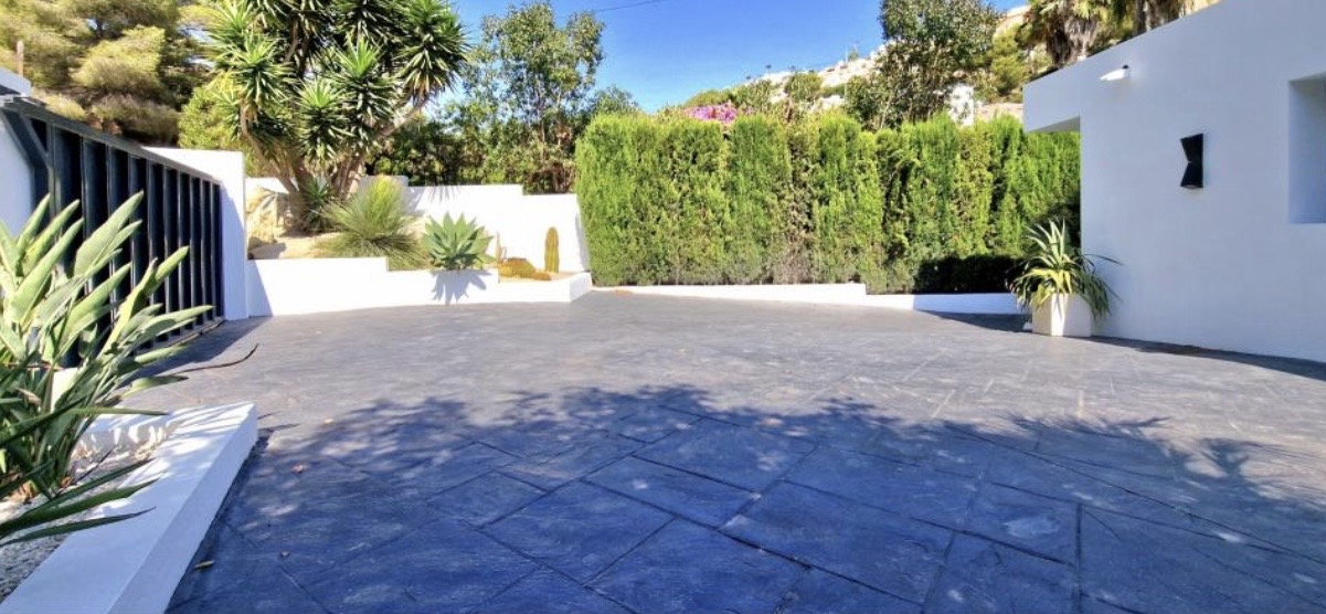 Spacious driveway featuring dark grey stamped concrete, bordered by white walls, a black metal gate, and a lush Mediterranean garden with yuccas.