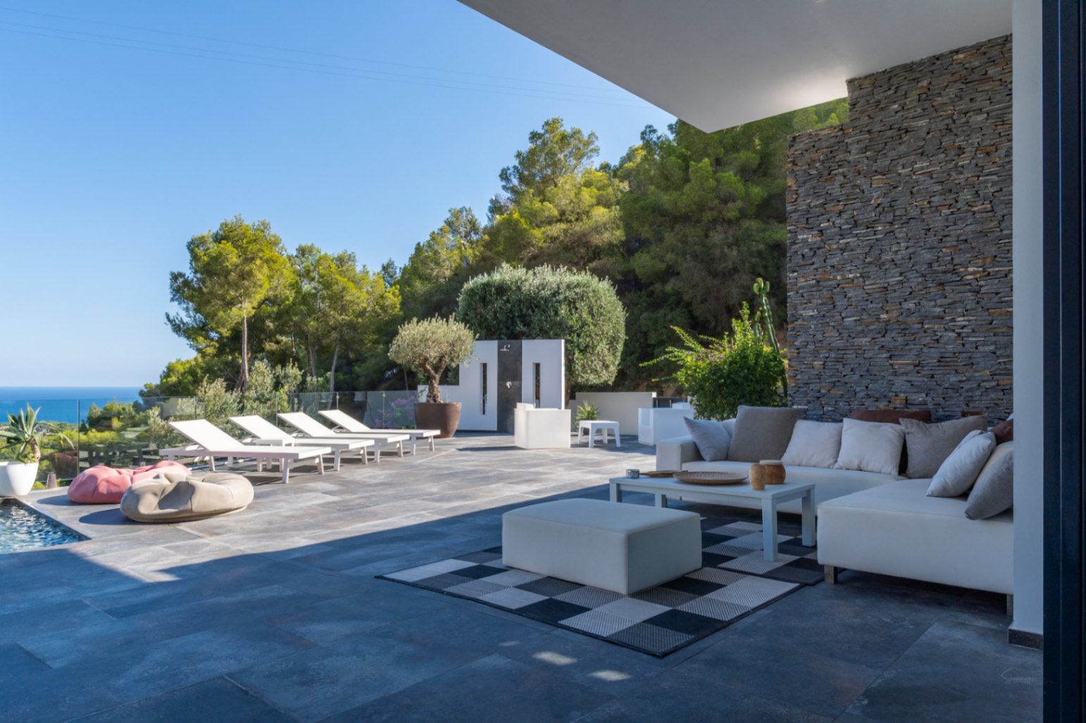 Spacious terrace featuring a white modular lounge area against a natural stone wall, leading to a sun deck with loungers and expansive sea views.