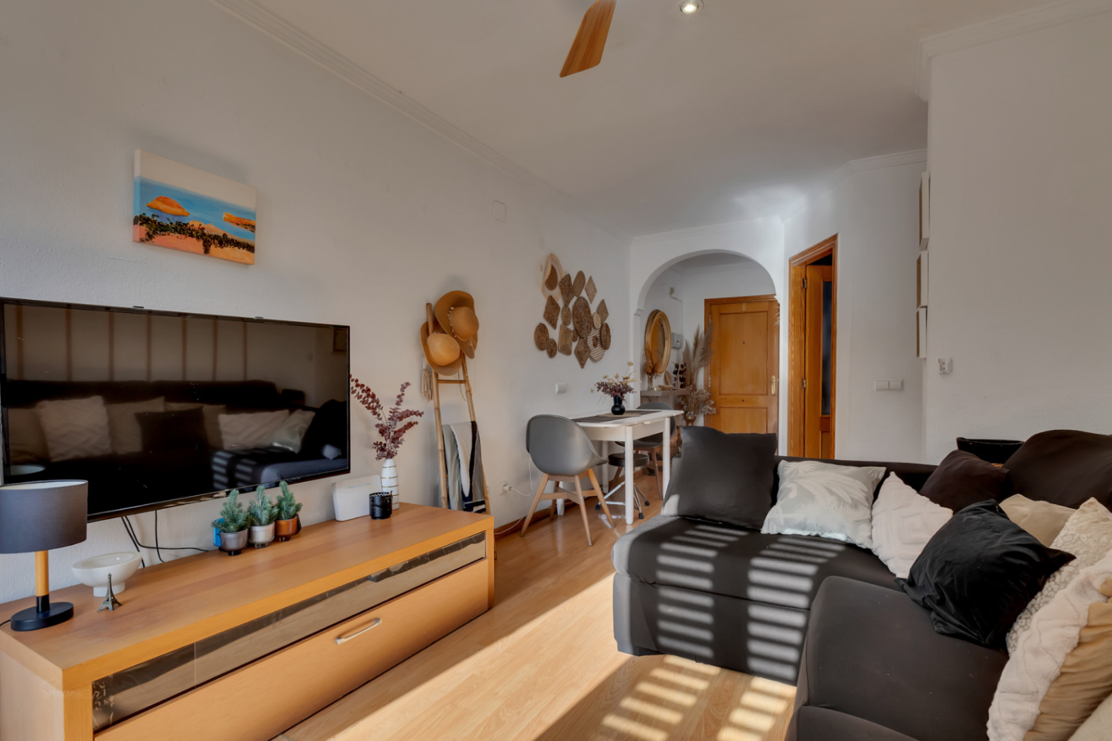 Bright living and dining area with wooden floors, a dark sofa, and a TV unit. Features a dining nook under a decorative arch and Mediterranean-style accents.