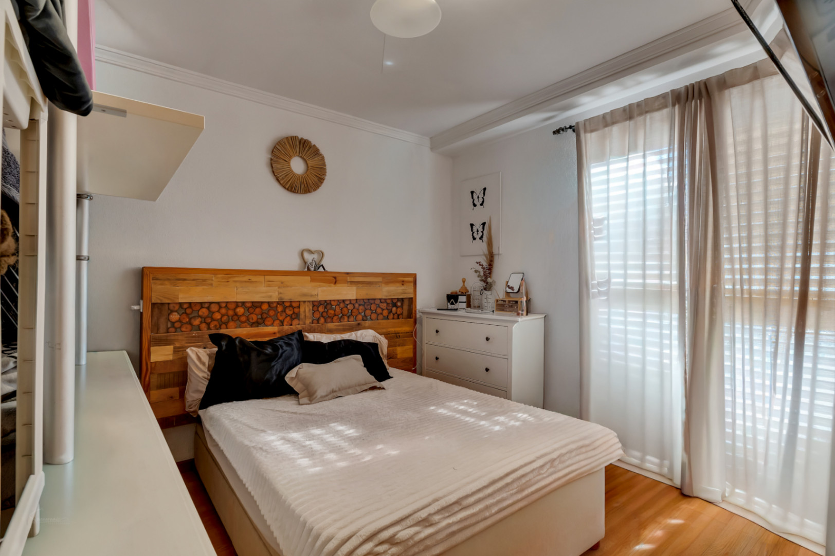 Master bedroom featuring a handcrafted rustic wooden headboard, laminate flooring, and a large window with sheer curtains for natural illumination.