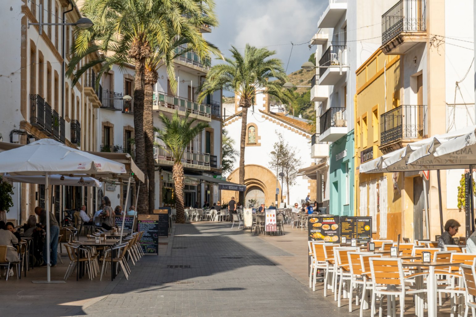 Pedestrian street in Jávea's historic center featuring restaurant terraces, palm trees, and traditional facades leading towards a stone chapel.