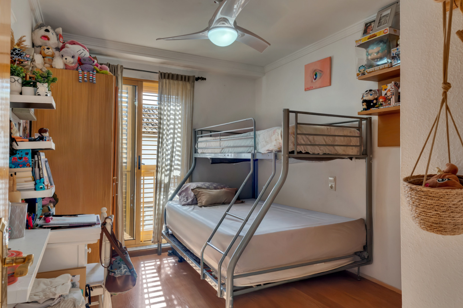 Secondary bedroom featuring a metal bunk bed, wooden flooring, and a ceiling fan. It offers direct balcony access through sliding doors with traditional wooden shutters.