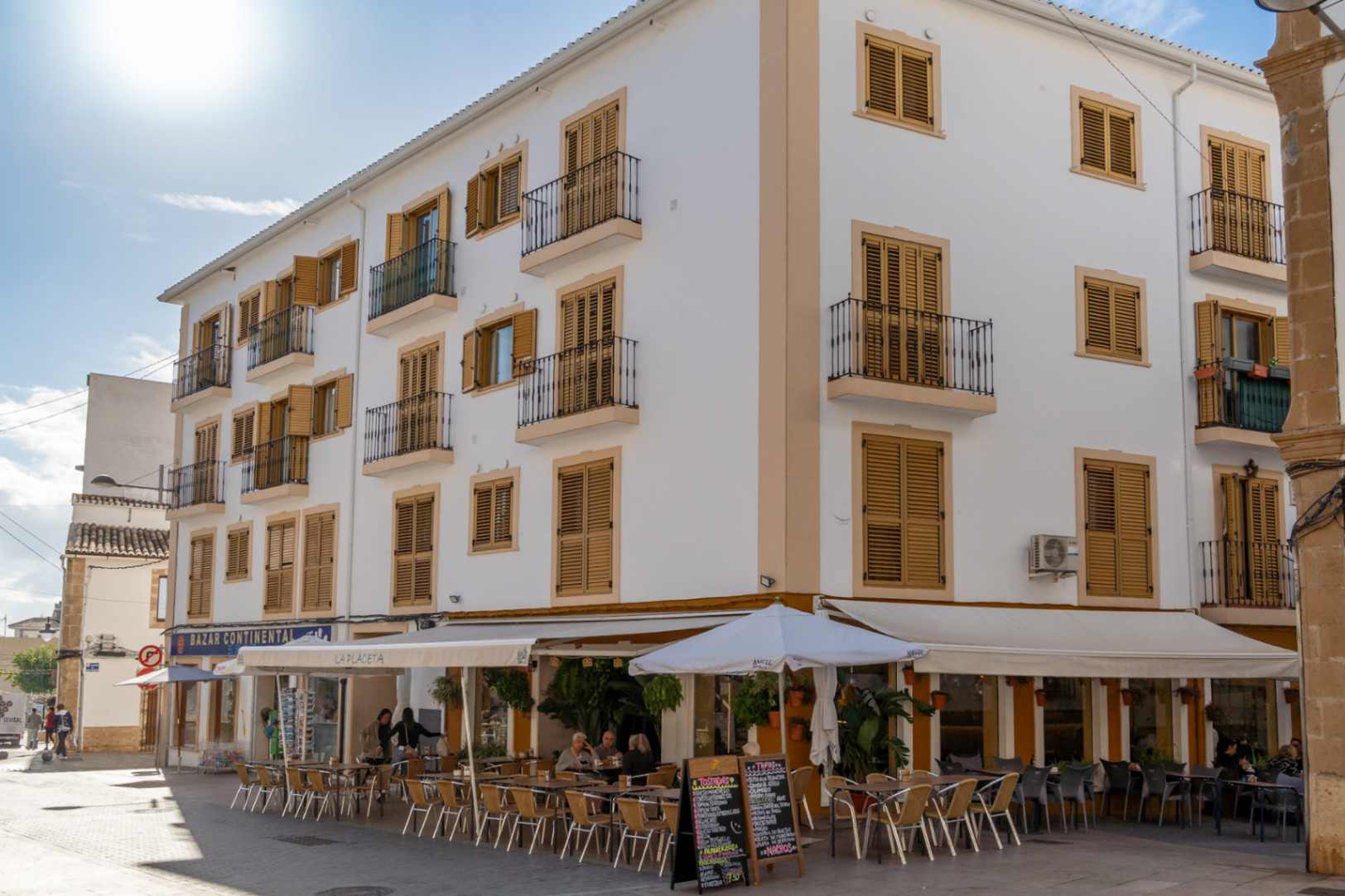 Traditional facade in the historic center featuring ochre wooden shutters, wrought-iron balconies, and a ground-floor cafe with outdoor seating in a pedestrian area.