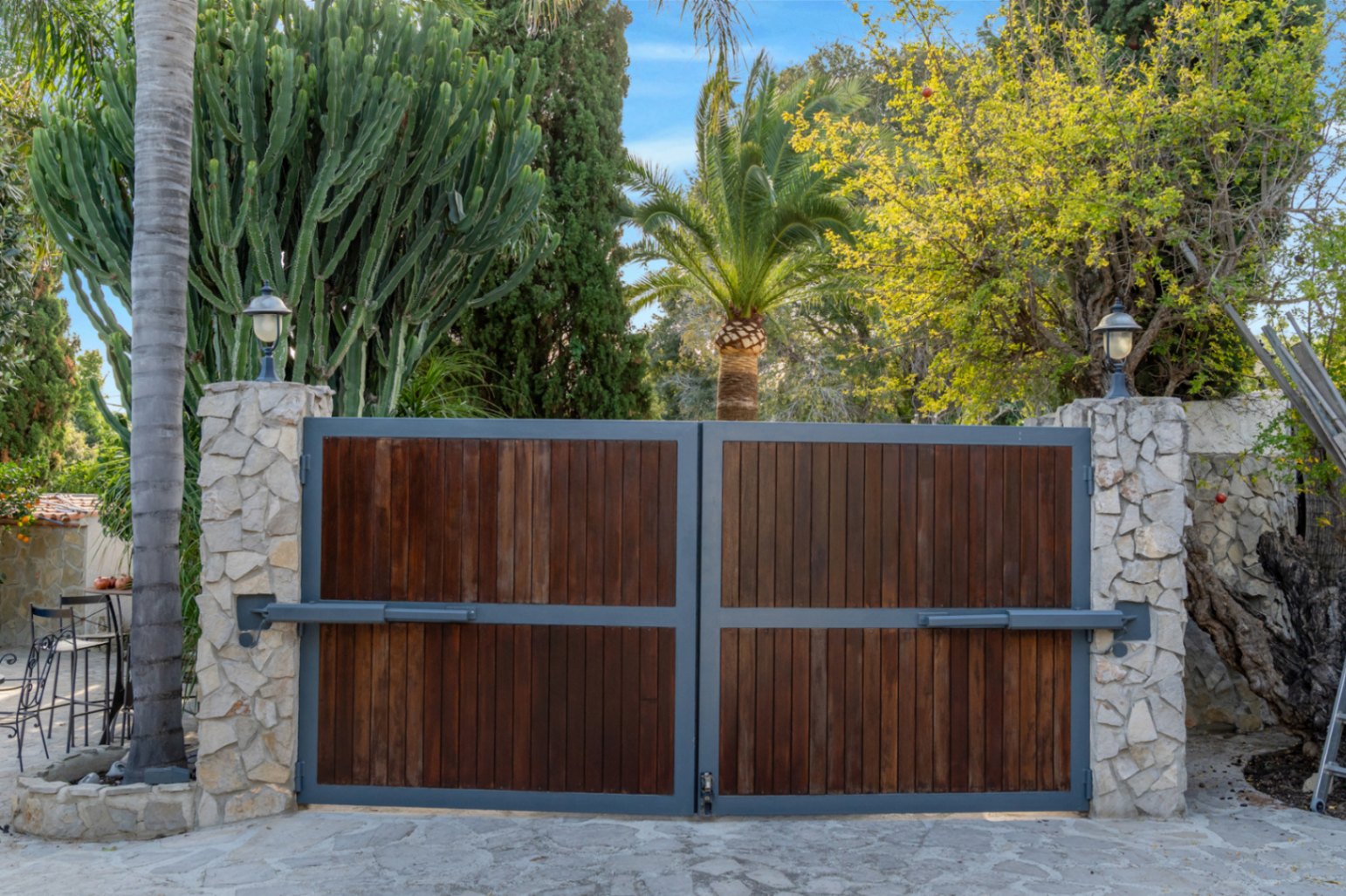 Automatic entrance gate featuring dark wood panels and grey metal framing, set between natural stone pillars and lush Mediterranean landscaping.