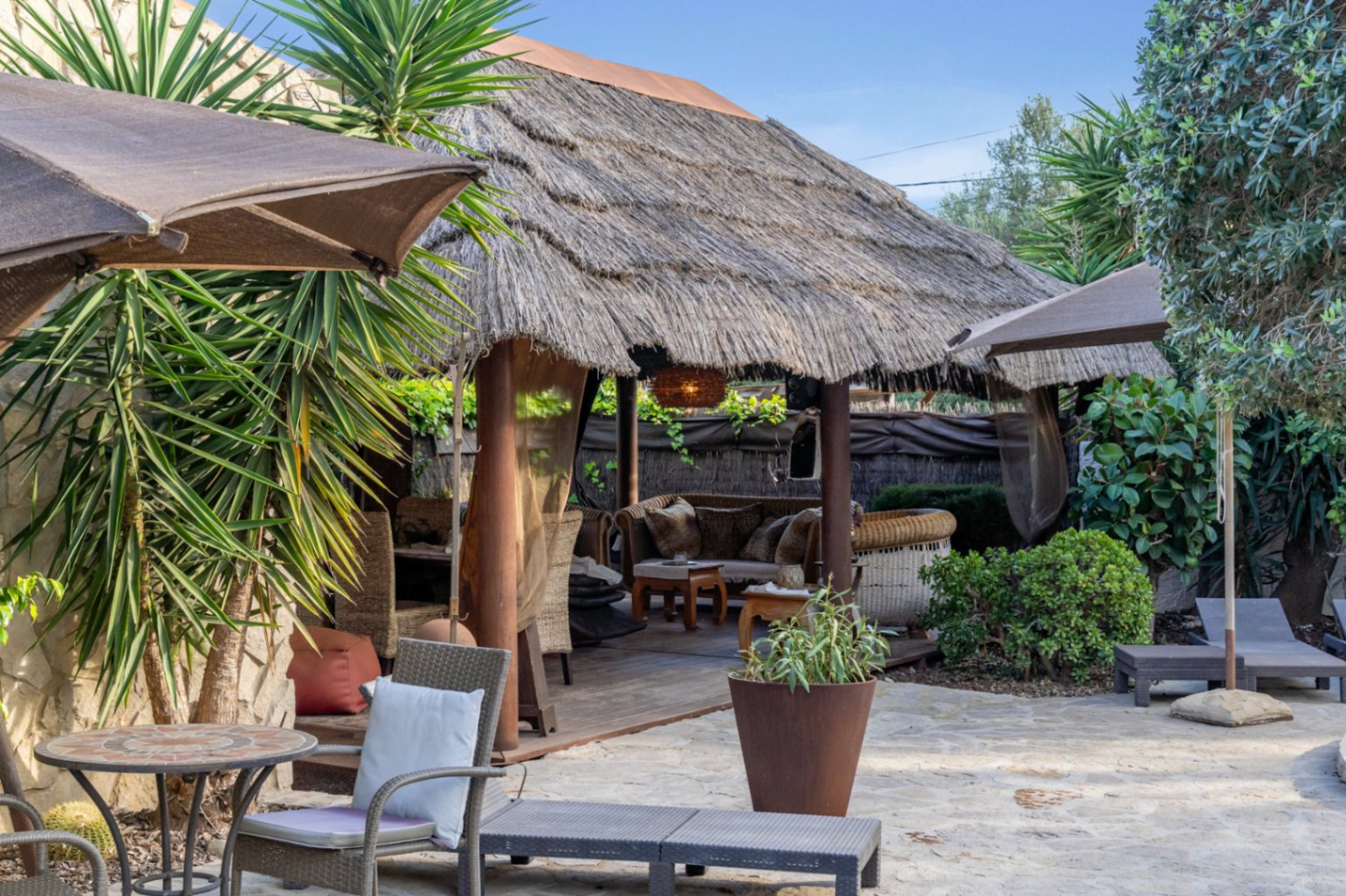 Balinese-style pergola featuring a thatched roof and wooden decking, set within a natural stone terrace with lounge seating and lush greenery.