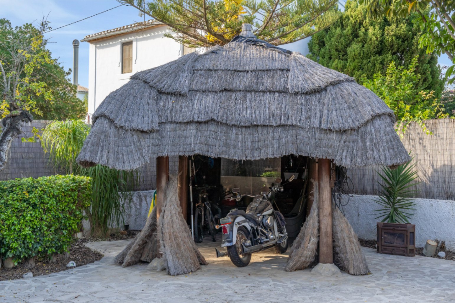 Balinese-style pergola with a thick thatched roof and wooden pillars, currently serving as a sheltered parking space for a motorcycle and bicycles in the garden.