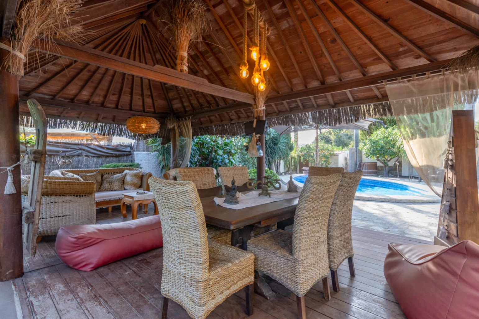 Balinese-style wooden pergola featuring an outdoor dining area with wicker chairs, rope lighting, and views of the private pool.