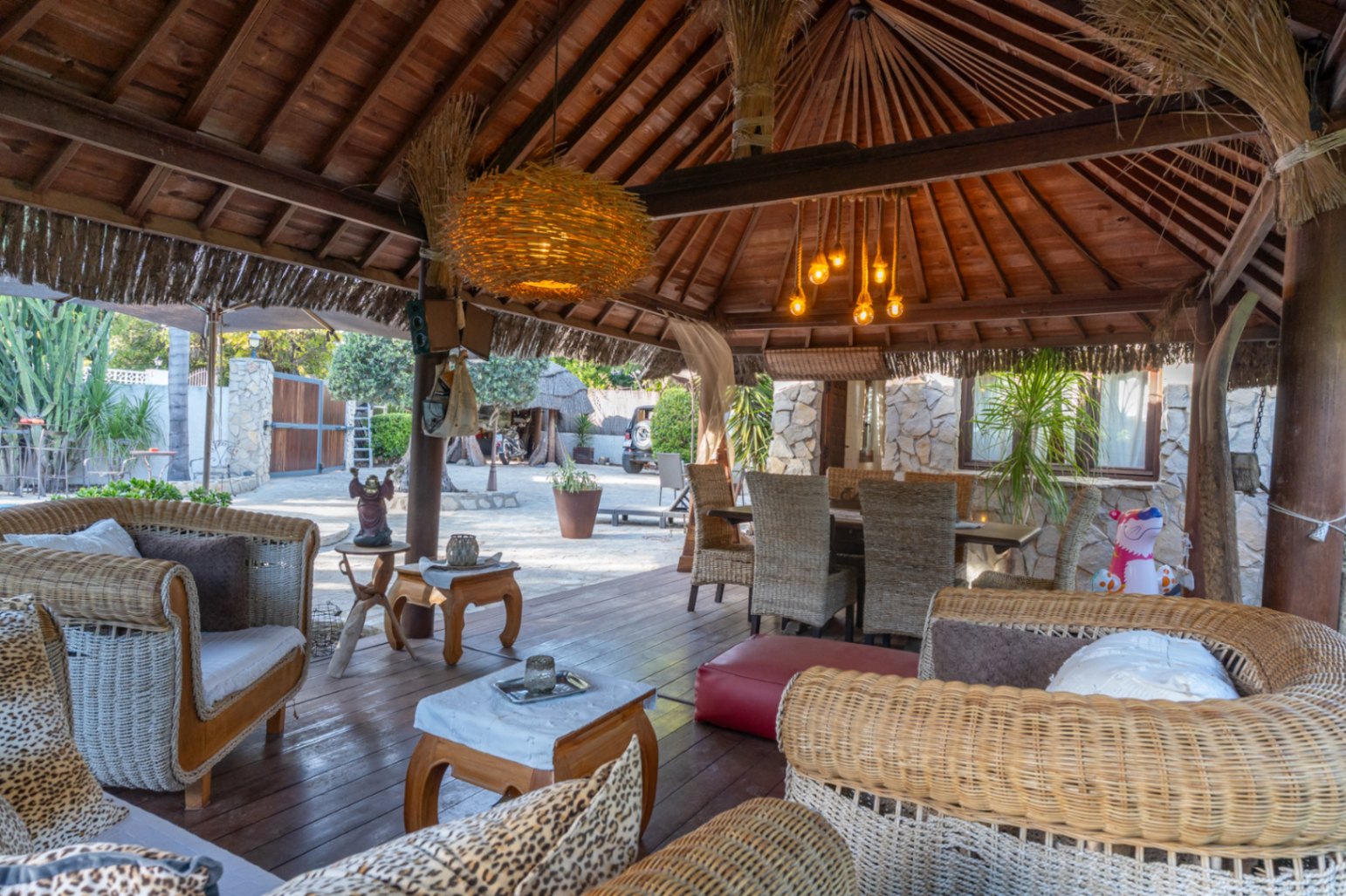 Balinese-style wooden pergola featuring exposed rafters, wicker lounge furniture, and an outdoor dining area set on dark wood decking.