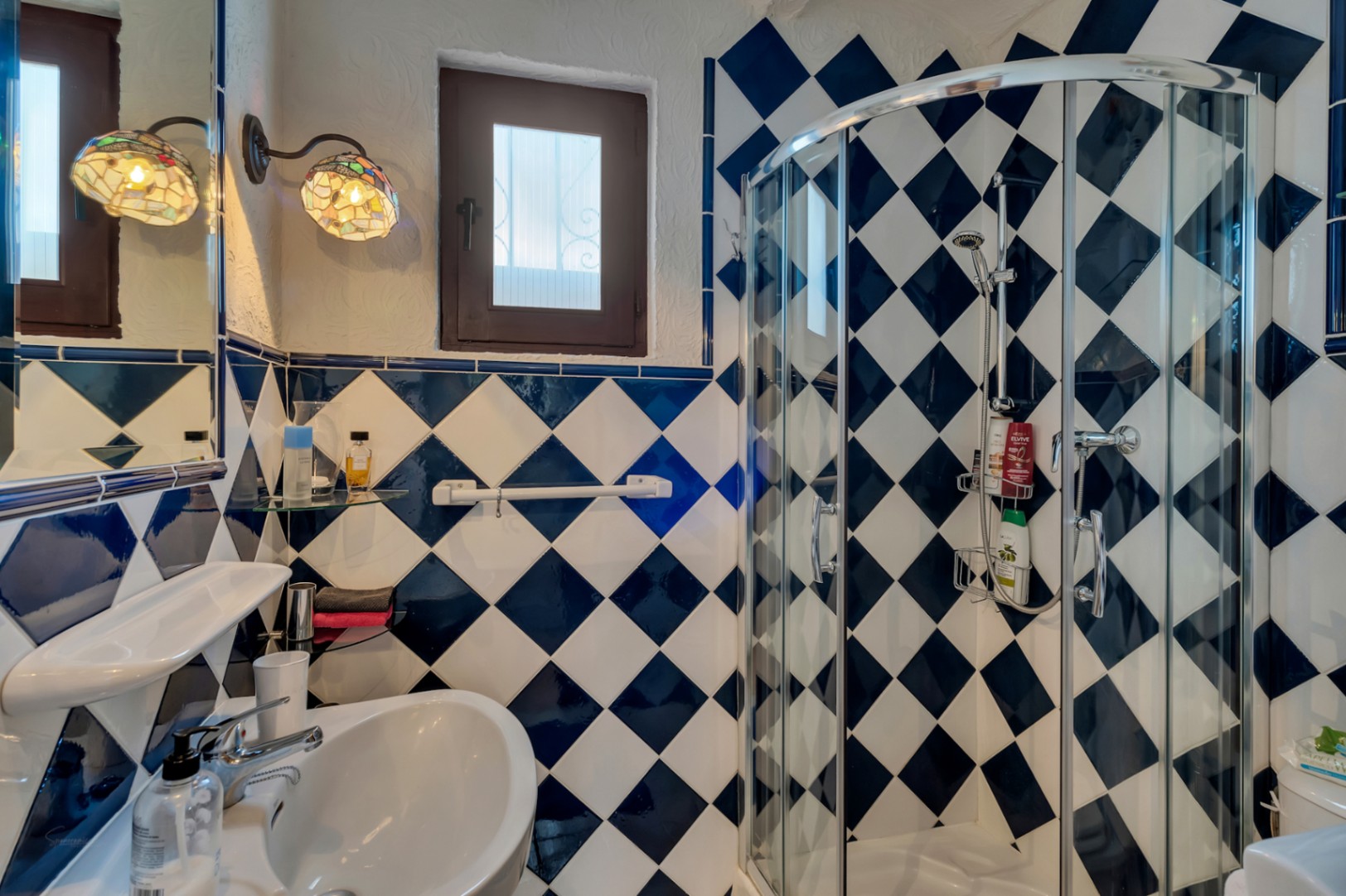 Bathroom featuring blue and white diamond-patterned tiling, a curved glass corner shower, and decorative Tiffany-style wall lamps.