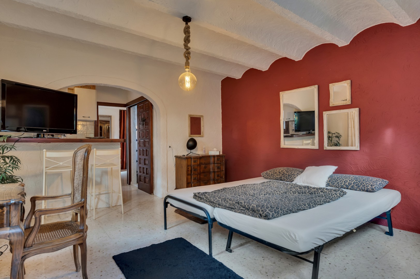 Bedroom featuring a red accent wall, vaulted ceilings, and terrazzo flooring. It includes an arched doorway to the living area and a rustic rope pendant light.