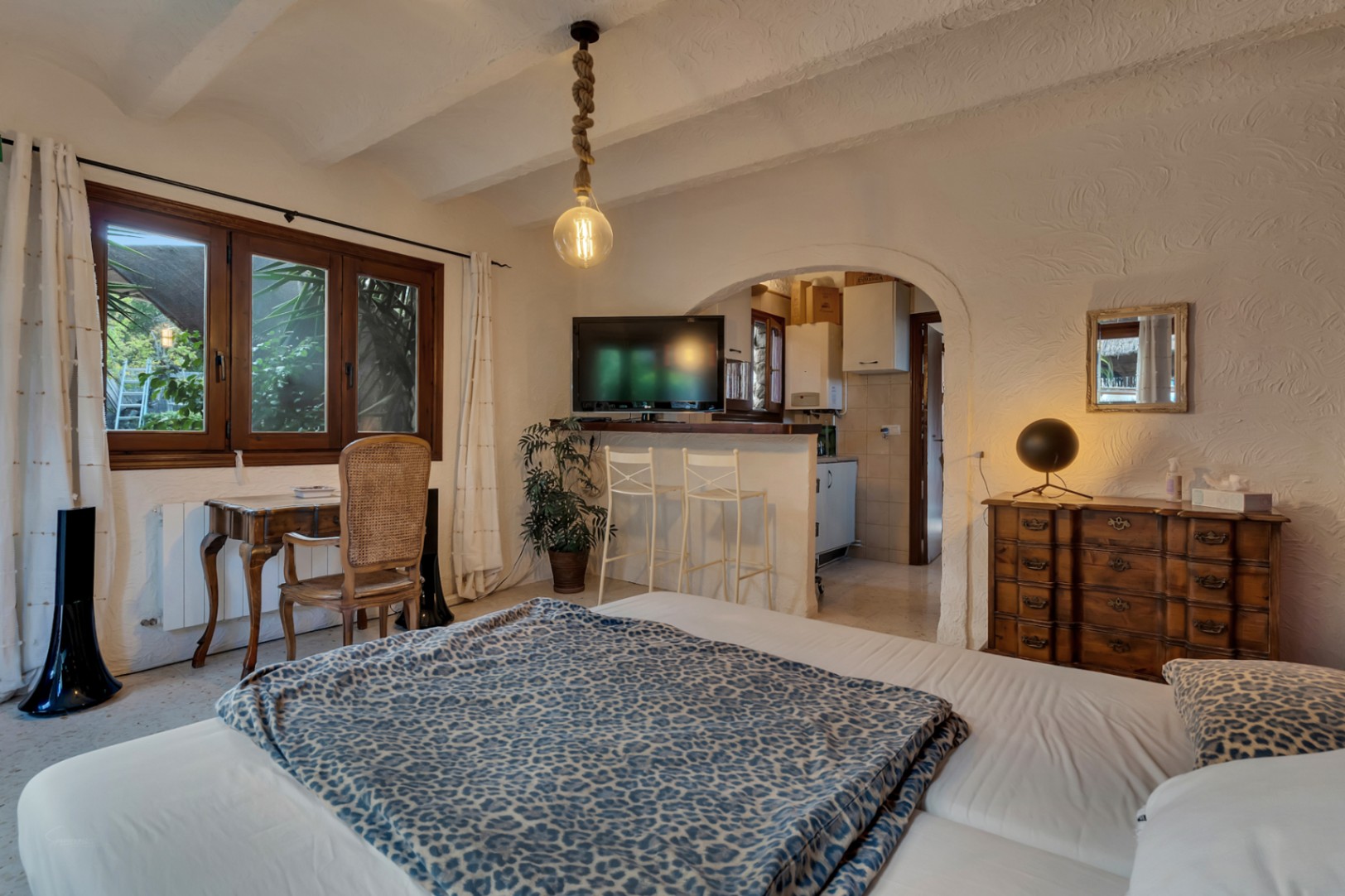 Bedroom featuring a wooden workspace, beamed ceiling, and an arched opening to a compact kitchenette with a breakfast bar and stools.