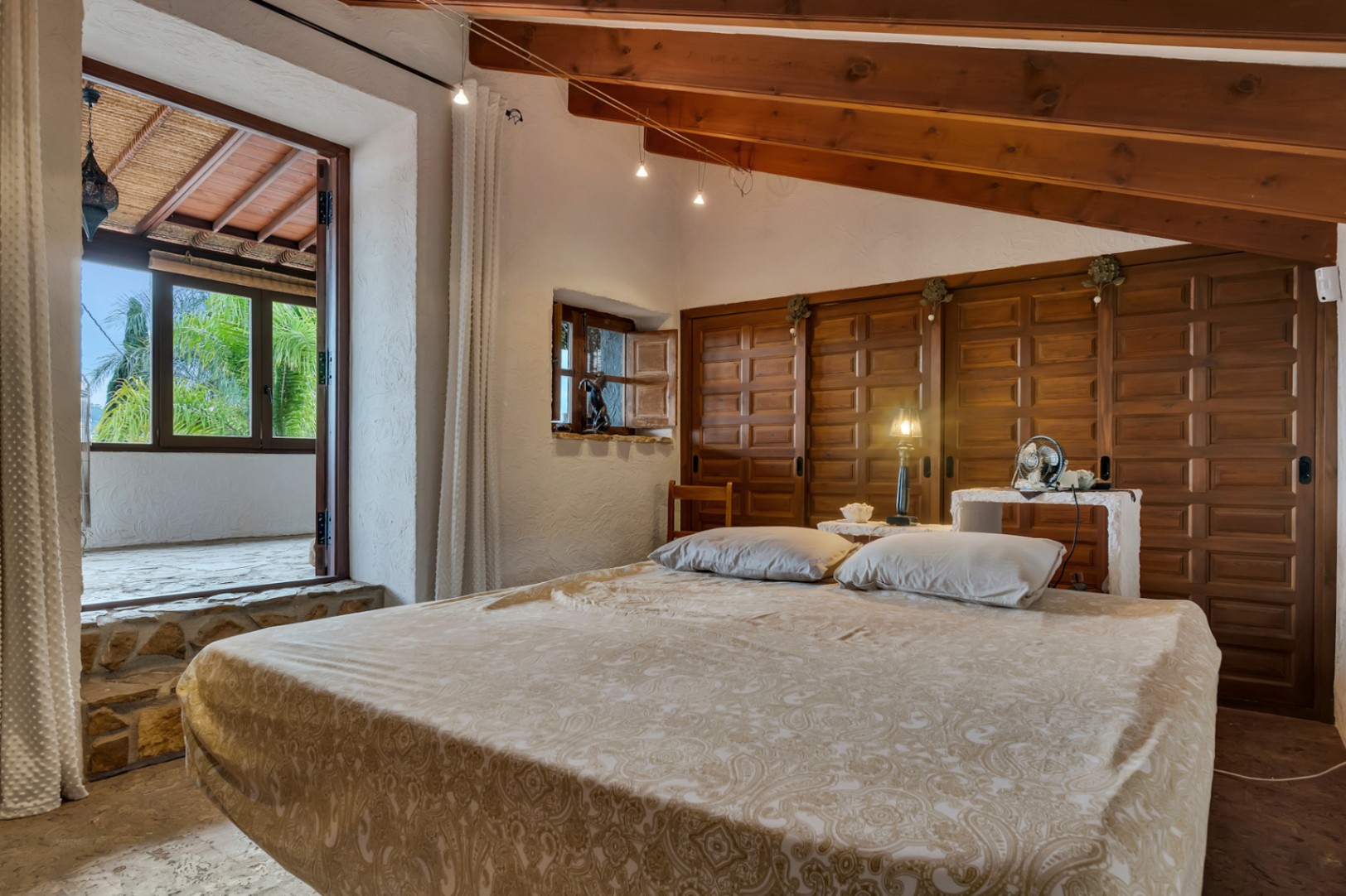 Bedroom featuring exposed wooden ceiling beams, traditional paneled wardrobes, and a stone step leading to a private covered terrace with garden views.