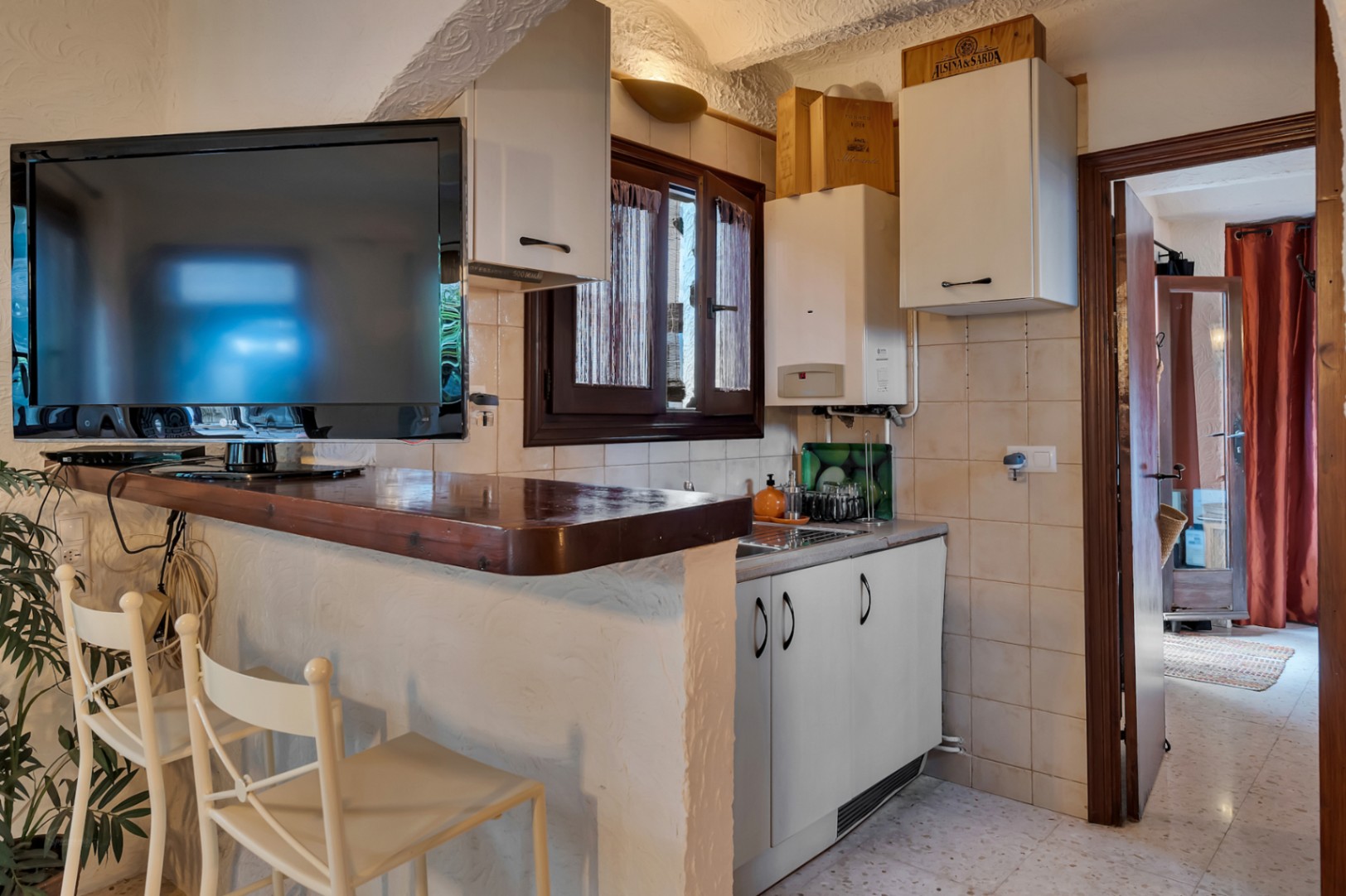 Compact kitchenette featuring a wooden breakfast bar, white cabinetry, and integrated television. The space includes textured plaster walls and a functional layout.