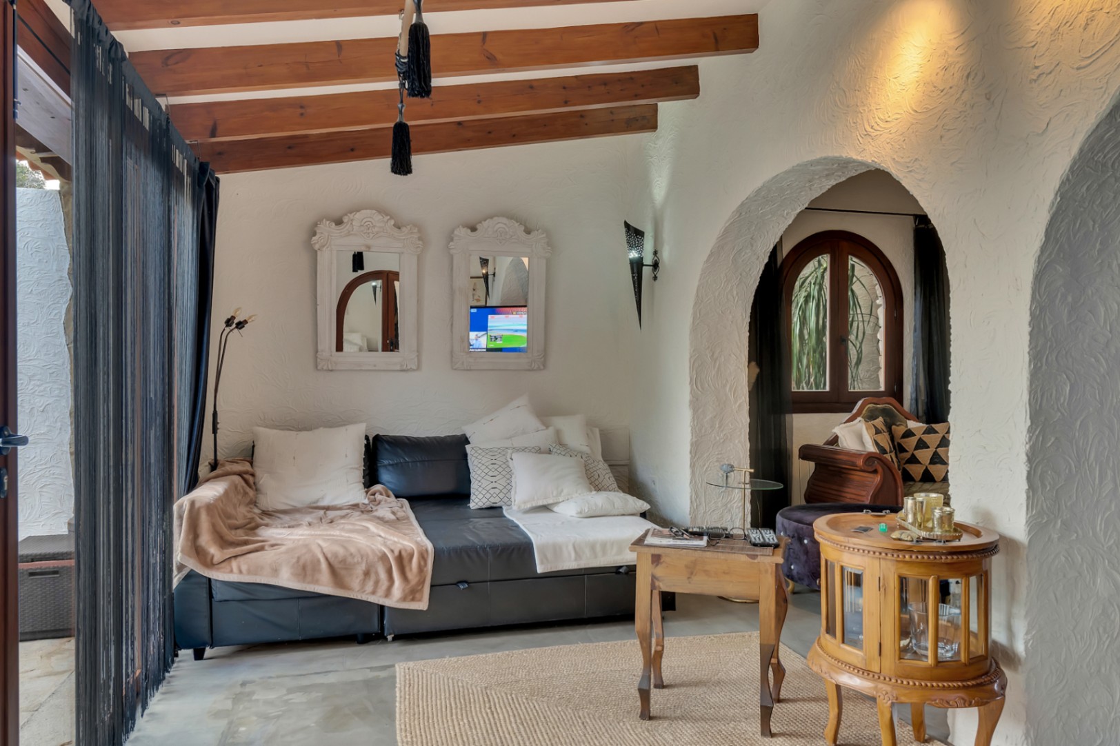 Cozy living area featuring exposed wooden ceiling beams, rustic textured walls, and traditional Mediterranean arches. Furnished with a leather sofa and vintage wooden accents.