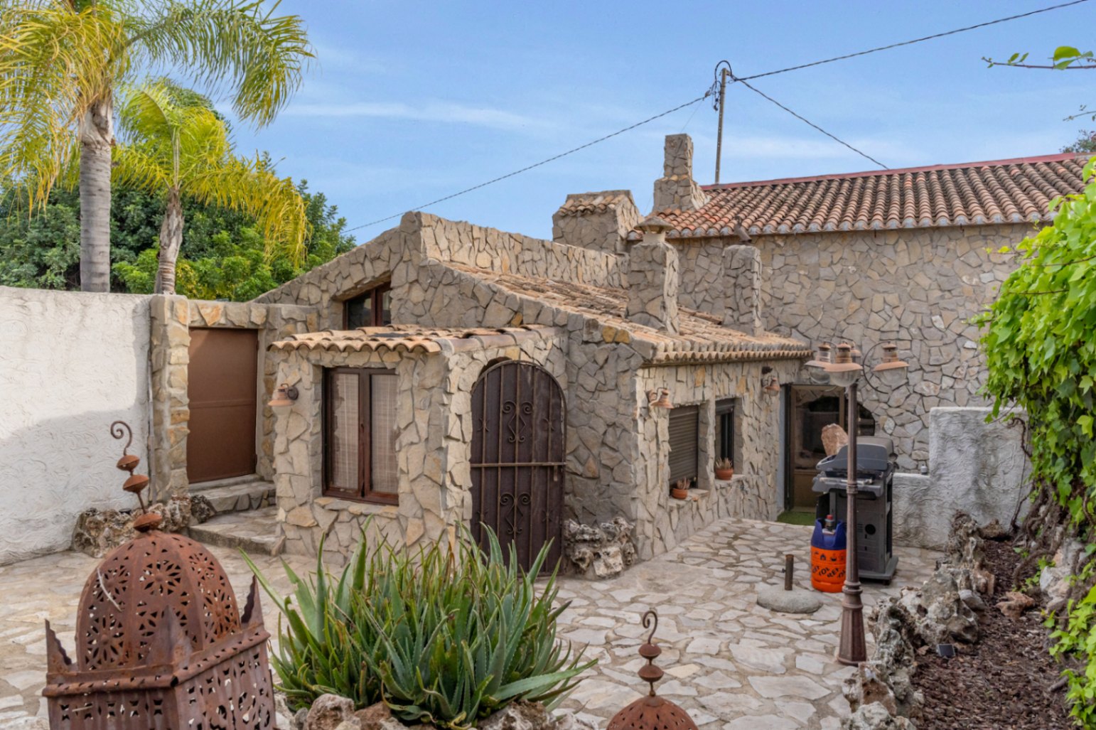 Detached guest apartment featuring natural stone walls, terracotta roof tiles, and a paved patio with an outdoor grill and Mediterranean vegetation.