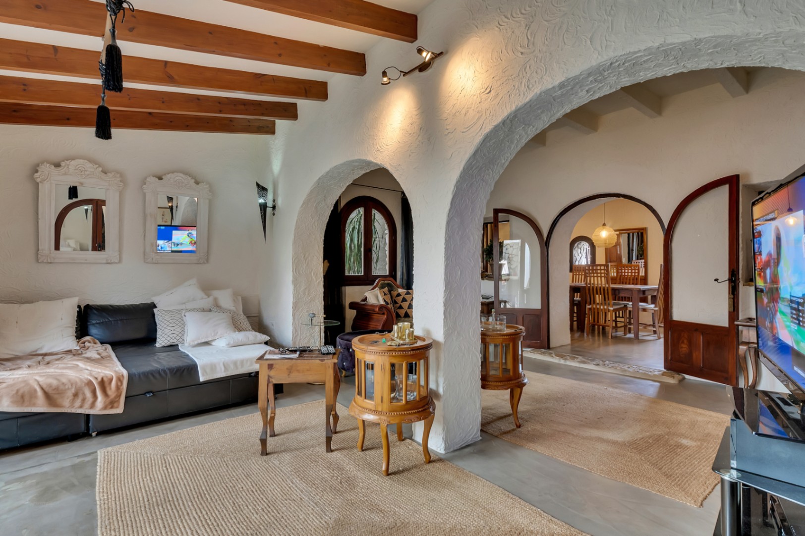 Living room with exposed wooden ceiling beams, traditional arches, and textured white walls, leading into the dining area through arched glass doors.