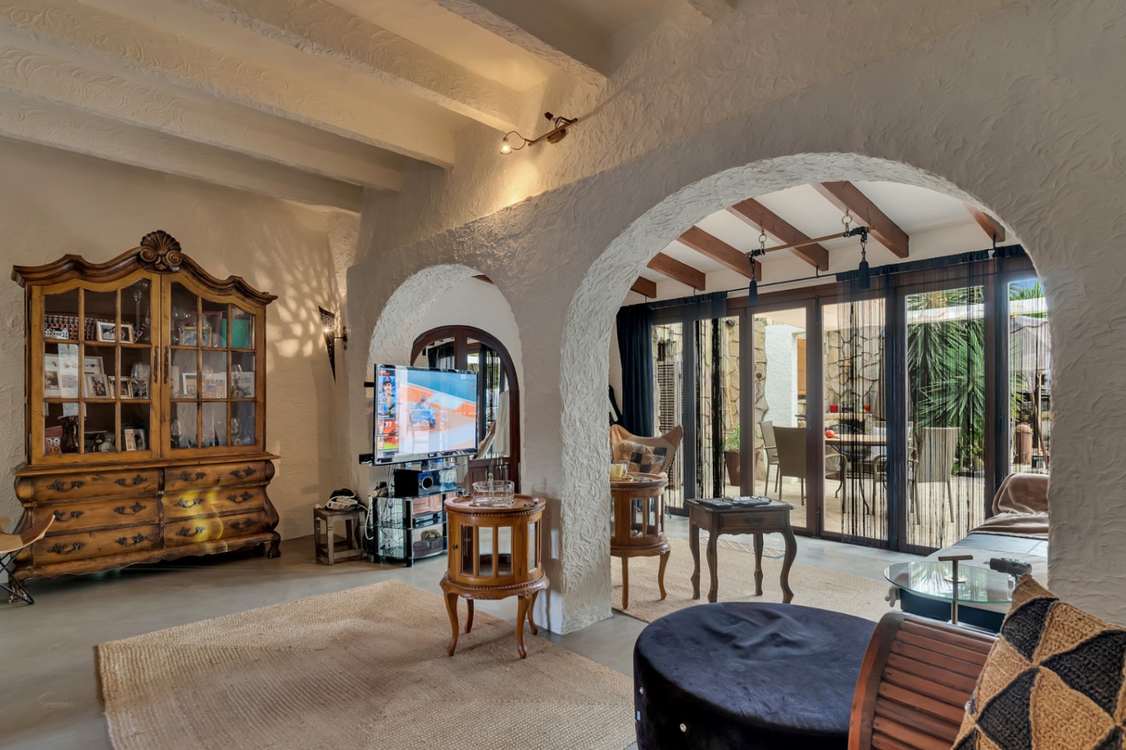 Mediterranean living area featuring textured white arches, exposed wooden beams, and a classic timber display cabinet. Large glass doors open directly to the patio.