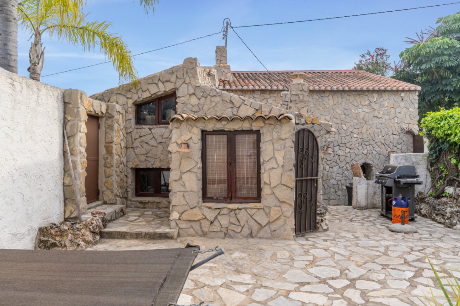Natural stone facade featuring dark wood window frames, a paved patio, and an outdoor barbecue area adjacent to the guest house.