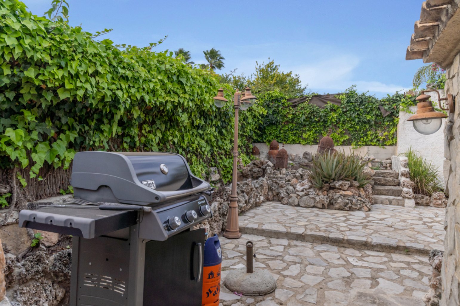 Outdoor barbecue area featuring a Weber gas grill, natural stone paving, and lush ivy-covered walls with rustic Mediterranean landscaping.