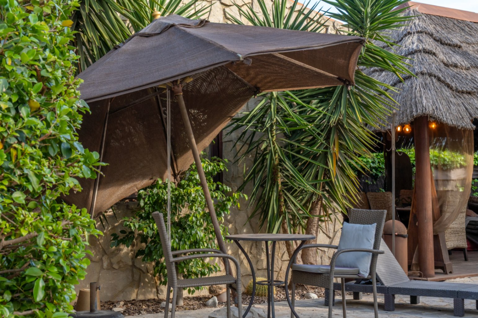 Outdoor seating area featuring a brown parasol, bistro table, and wicker chairs, set against a natural stone wall and a thatched Balinese pergola.