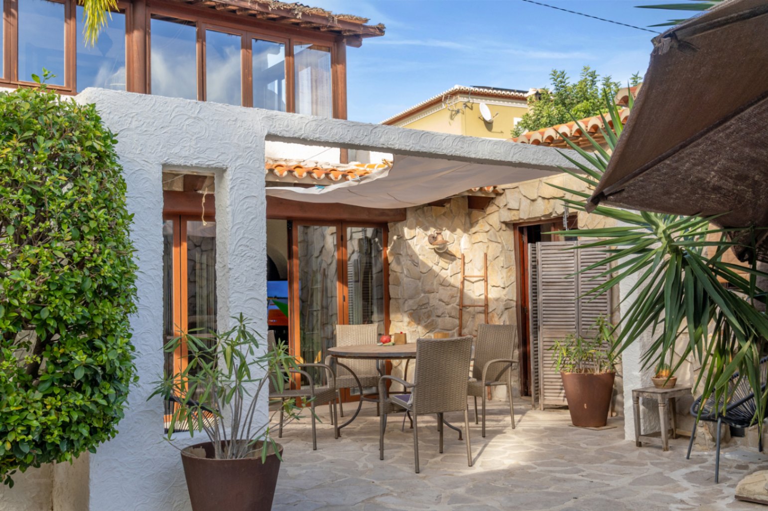 Outdoor terrace featuring natural stone paving, a dining set under a sail awning, and a facade blending rustic stone masonry with wooden frames.