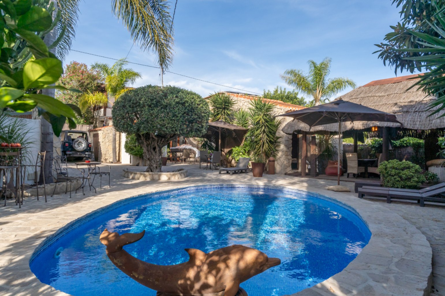 Oval swimming pool framed by natural stone paving, featuring a Balinese-style thatched pergola, mature palm trees, and integrated lounge areas.