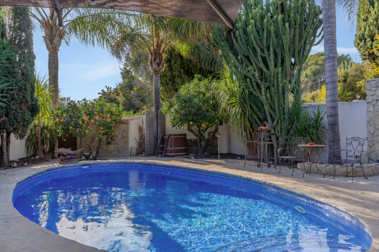 Private swimming pool with blue mosaic tiling, surrounded by mature Mediterranean gardens with palm trees, citrus, and a wrought iron seating area on stone paving.