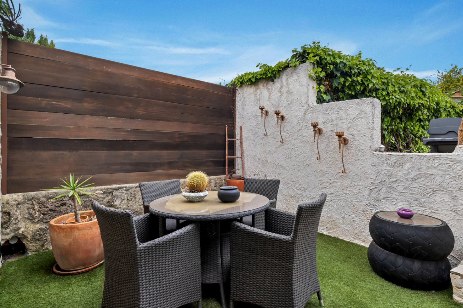 Private terrace with artificial grass, a circular dining set in dark wicker, and walls featuring textured plaster, wood panels, and iron accents.