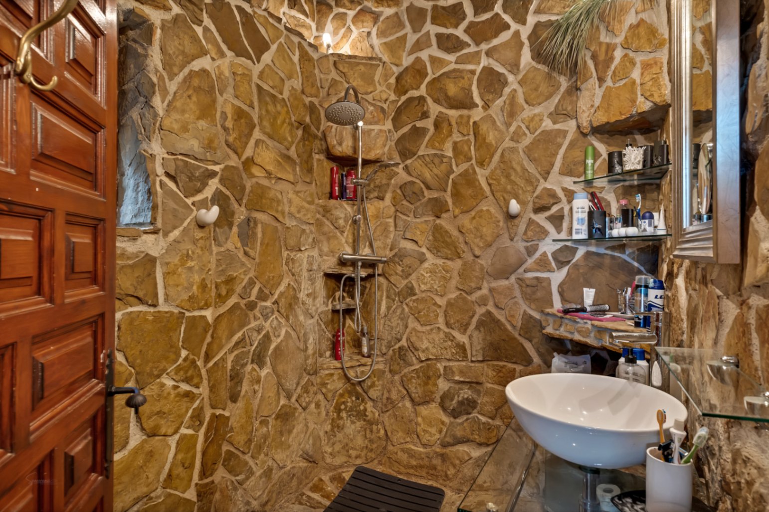 Rustic bathroom featuring irregular natural stone walls, a walk-in shower with stainless steel fixtures, and a modern vessel sink on a glass vanity.