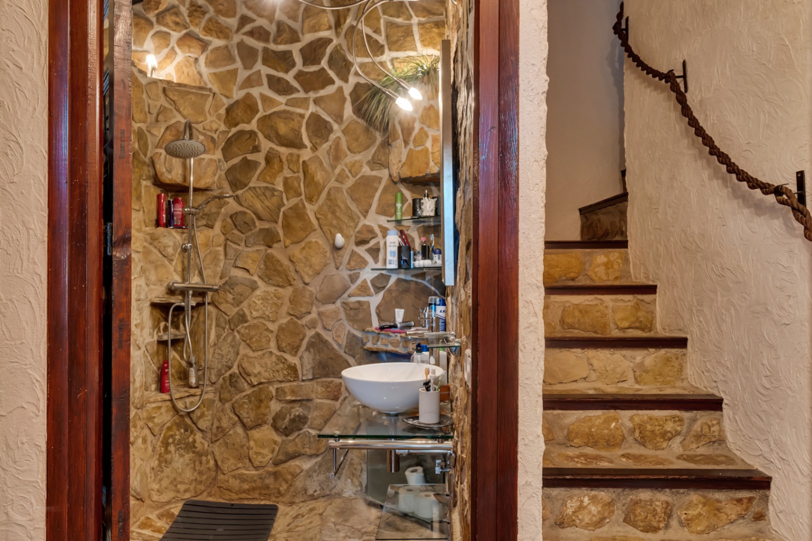 Rustic bathroom featuring natural stone walls, a walk-in shower, and a vessel sink, situated next to a stone staircase with a rope handrail.