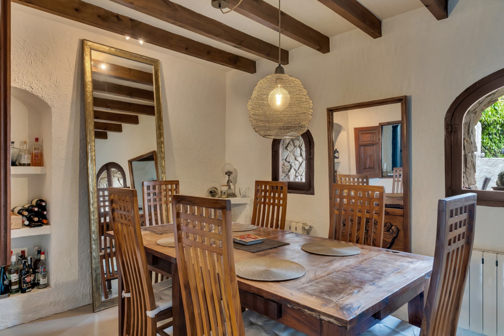 Rustic dining area featuring exposed wooden beams, a solid wood table, and high-back chairs. Integrated wall niches, large mirrors, and a woven pendant light complete the space.