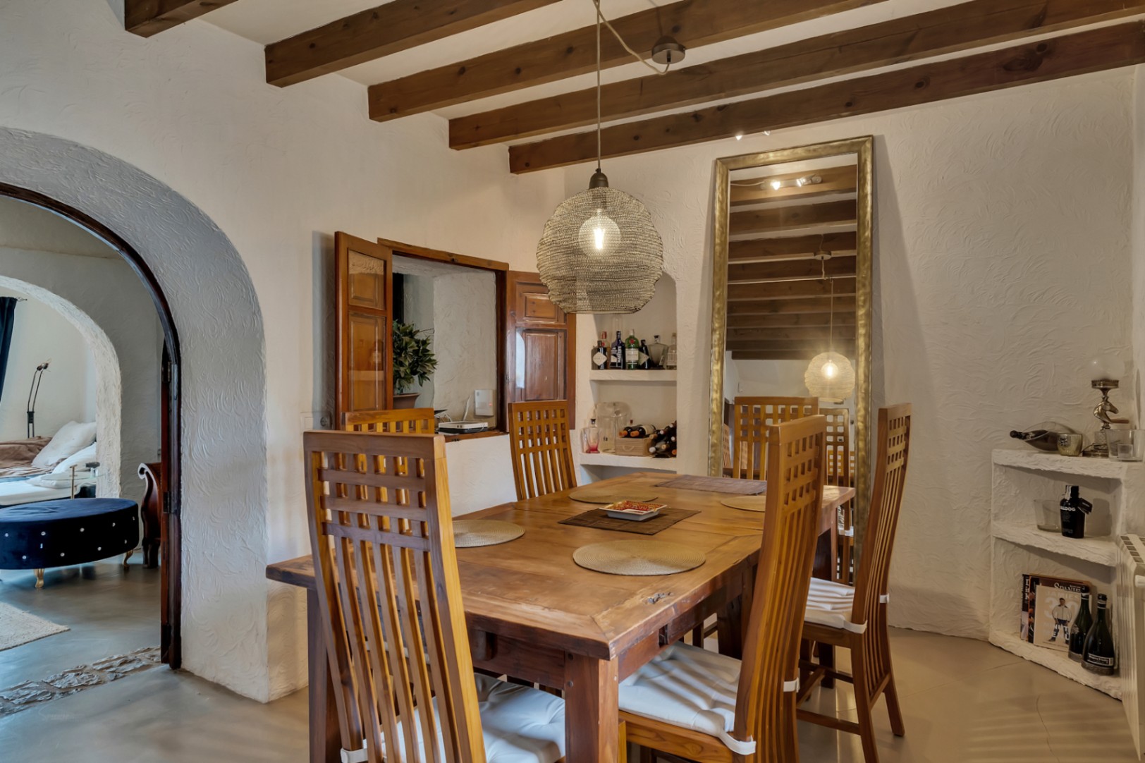 Rustic dining area featuring exposed wooden beams, a solid wood table, and high-back chairs. The space includes a serving hatch with wooden shutters and an elegant arched doorway.
