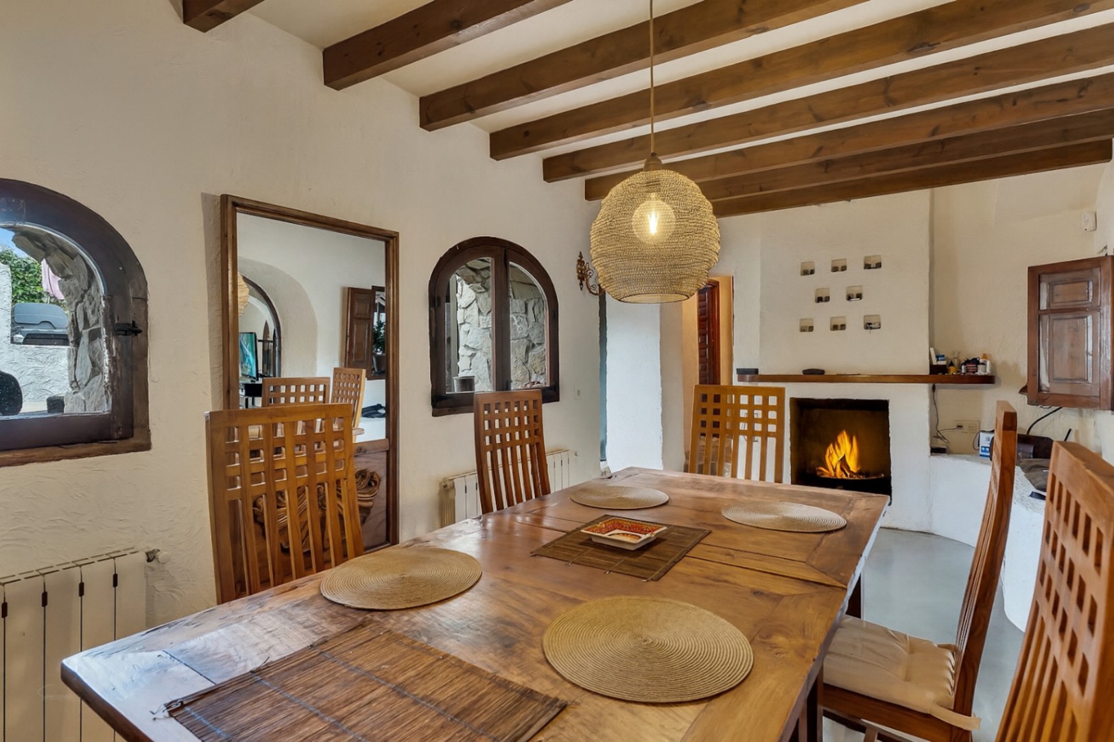 Rustic dining area featuring exposed wooden ceiling beams, a solid wood table, and a built-in fireplace. Arched windows and a woven pendant light enhance the atmosphere.