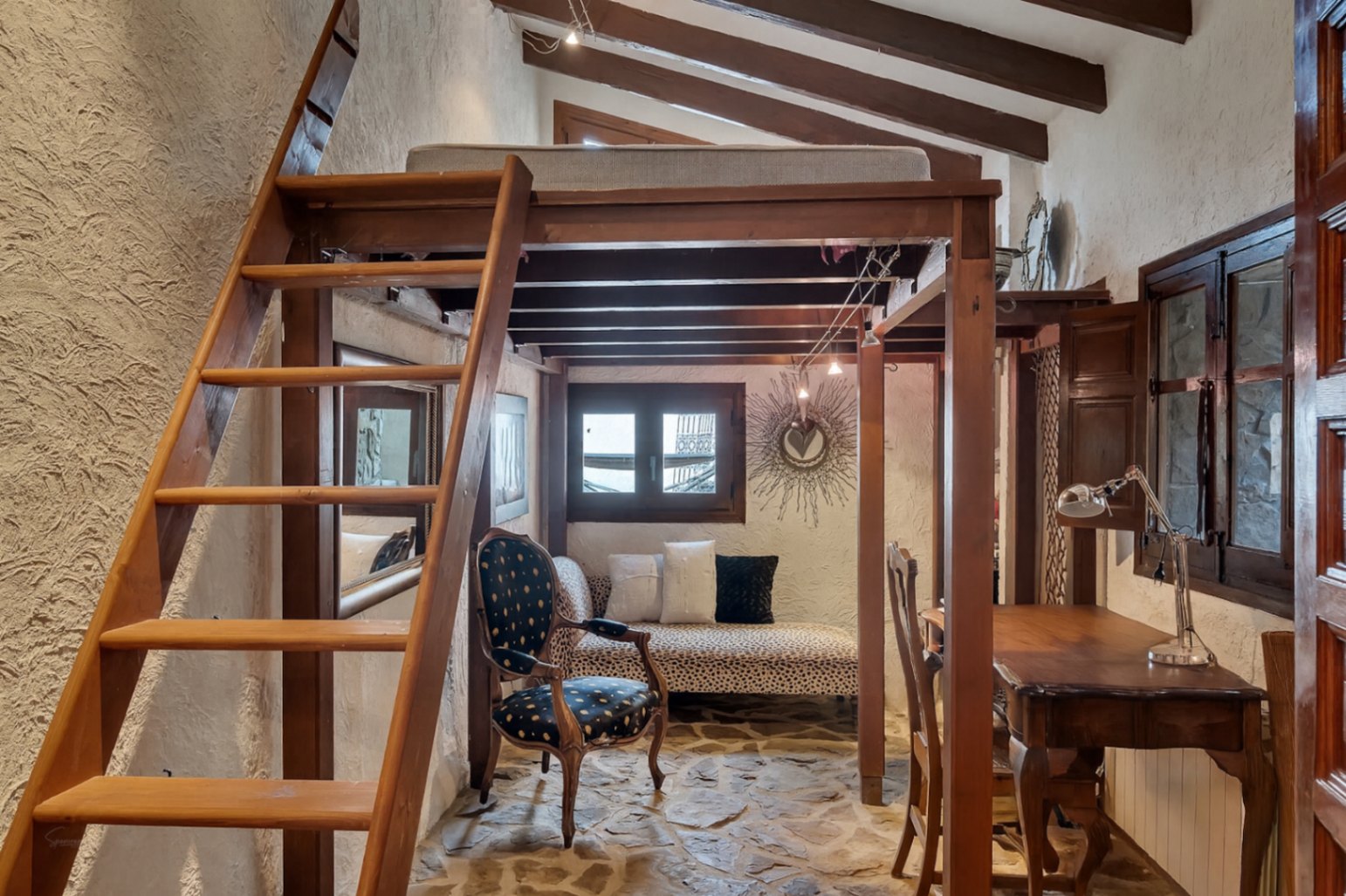 Rustic room featuring a wooden mezzanine bed, natural stone flooring, and exposed beams. The space includes a lower seating area and desk under the loft structure.