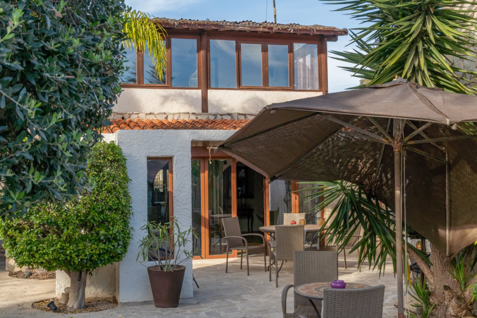 Villa exterior featuring a glazed upper terrace, stone-paved patio with outdoor dining furniture, and lush Mediterranean landscaping.