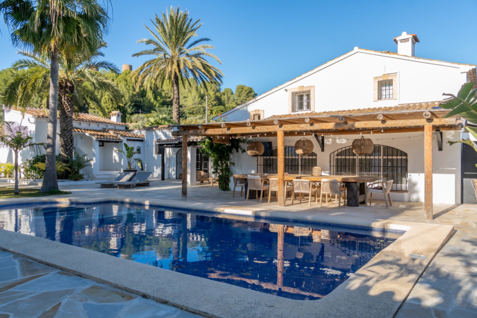 Exterior view featuring a rectangular swimming pool and a wooden pergola. The outdoor dining area includes a large table, wicker lamps, and traditional Mediterranean architecture.