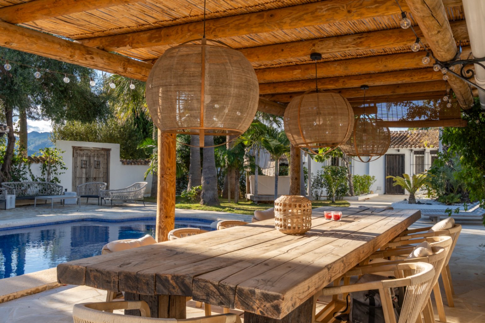 Outdoor dining area under a wooden beam and reed pergola, featuring a rustic table, natural fiber pendant lights, and views of the pool and Mediterranean garden.