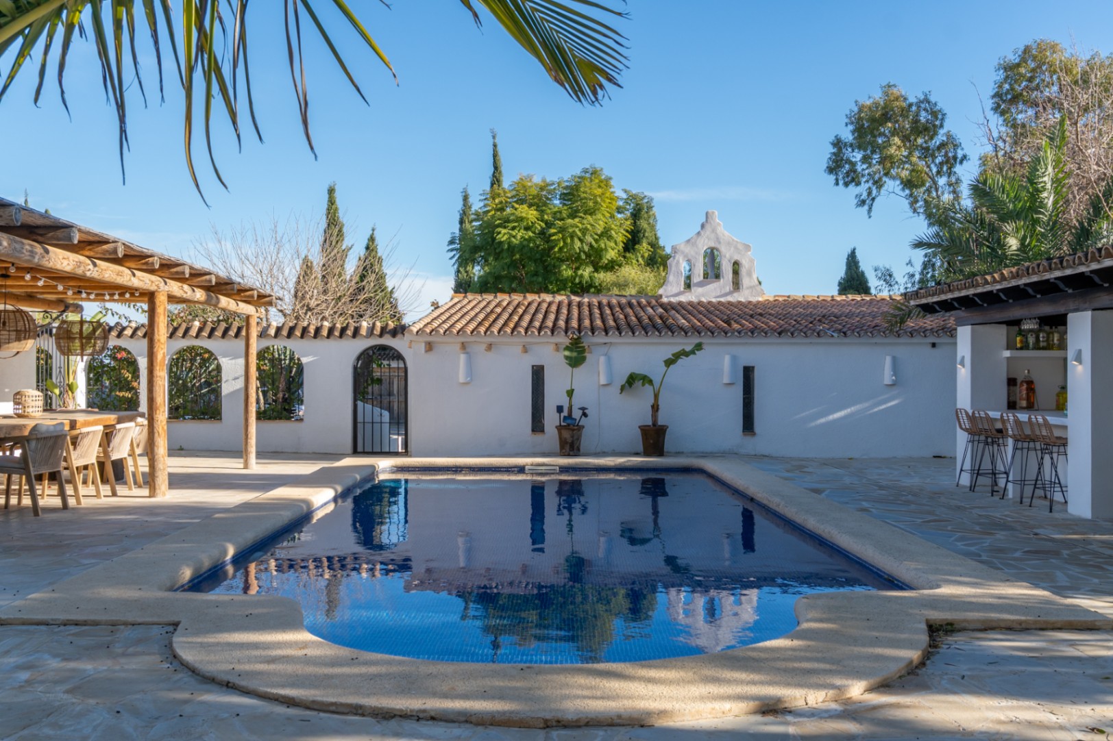 Outdoor pool area featuring blue tiling, a stone terrace, a wooden pergola for dining, and a summer bar with seating under a clear sky.