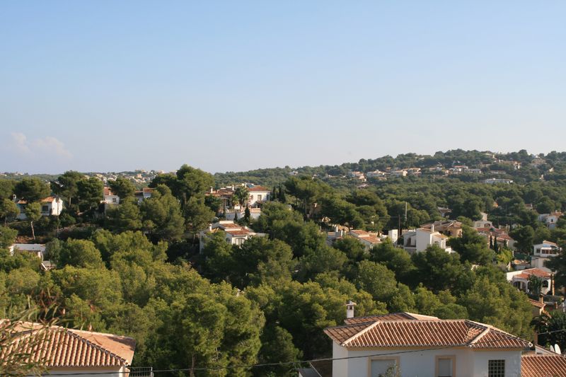 Panoramic view from the plot overlooking a residential area integrated into a Mediterranean pine forest, featuring traditional villas with terracotta roofs.