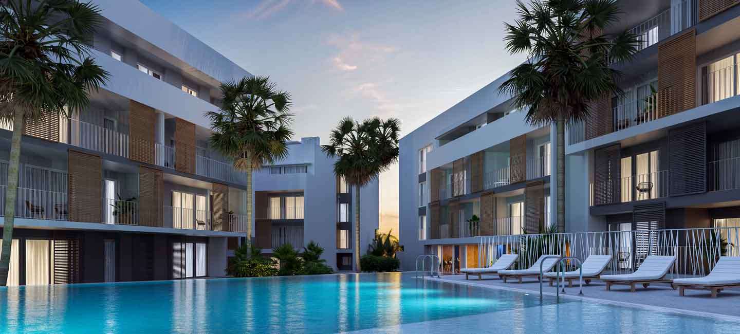 Exterior view of the modern apartment complex featuring a large communal pool, white facades with wooden privacy screens, and palm trees at dusk.